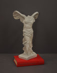 photo of plaster cast sculpture of winged, headless female figure with flowing drapery atop a red book on a gray background