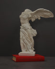 photo of plaster cast sculpture of winged, headless female figure with flowing drapery atop a red book on a gray background