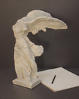 photo of plaster cast sculpture of winged, headless female figure with flowing drapery next to a pen and pad on a gray background