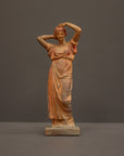 photo of plaster cast statue of a female in pink-orange tinted robes with her arms raised to her head against gray background