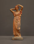photo of plaster cast statue of a female in pink-orange tinted robes with her arms raised to her head against gray background