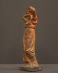 photo of plaster cast statue of a female in pink-orange tinted robes with her arms raised to her head against gray background