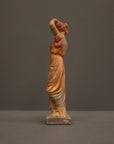 photo of plaster cast statue of a female in pink-orange tinted robes with her arms raised to her head against gray background