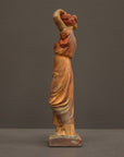 photo of plaster cast statue of a female in pink-orange tinted robes with her arms raised to her head against gray background