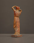 photo of plaster cast statue of a female in pink-orange tinted robes with her arms raised to her head against gray background