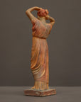 photo of plaster cast statue of a female in pink-orange tinted robes with her arms raised to her head against gray background