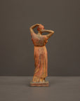 photo of plaster cast statue of a female in pink-orange tinted robes with her arms raised to her head against gray background