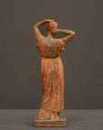 photo of plaster cast statue of a female in pink-orange tinted robes with her arms raised to her head against gray background
