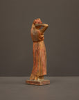 photo of plaster cast statue of a female in pink-orange tinted robes with her arms raised to her head against gray background