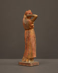 photo of plaster cast statue of a female in pink-orange tinted robes with her arms raised to her head against gray background