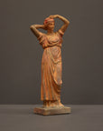 photo of plaster cast statue of a female in pink-orange tinted robes with her arms raised to her head against gray background