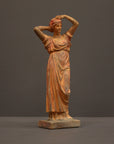 photo of plaster cast statue of a female in pink-orange tinted robes with her arms raised to her head against gray background