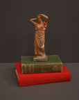 photo of plaster cast statue of a female in pink-orange tinted robes with her arms raised to her head atop green and red books against gray background