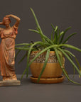photo of plaster cast statue of a female in pink-orange tinted robes with her arms raised to her head beside potted plant against gray background