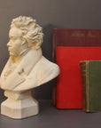 photo of plaster cast sculpture of male bust of Ludwig van Beethoven in dress coat and ruffled necktie set beside green and red books on gray background