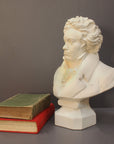 photo of plaster cast sculpture of male bust of Ludwig van Beethoven in dress coat and ruffled necktie set beside green and red books on gray background