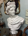 photo of plaster cast sculpture bust of man, namely the god Apollo, with hair piled high in the front and a broach near his neck holding robes and set on a wooden table against a blue pinstriped wall surrounded by antiques