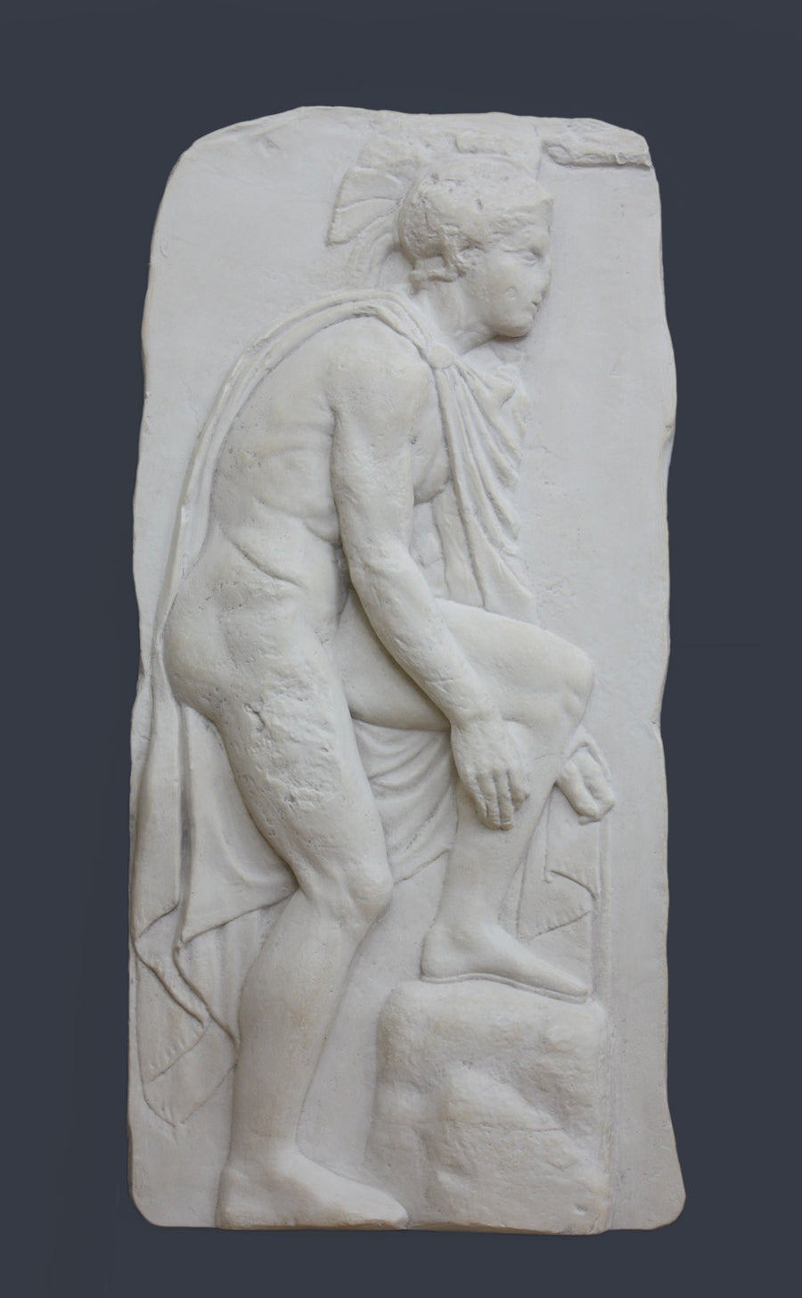photo of off-white plaster cast relief sculpture of male nude with cloak bending to tie his sandal against gray background