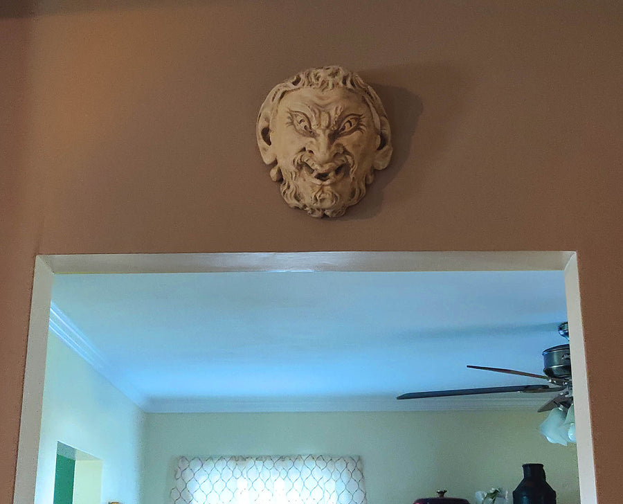 photo of plaster cast sculpture of grotesque faun face, affixed to beige wall above doorway to room with light walls and ceiling fan