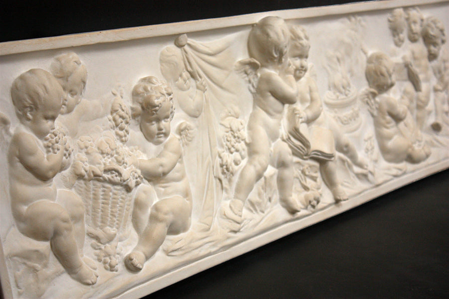 side view photo of white plaster cast with warm tones of relief sculpture of several cupids talking together with books and baskets of fruit on a black background