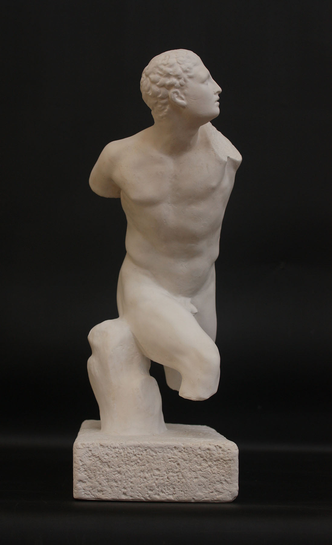photo of white plaster cast sculpture of male figure in active pose without arms or lower legs on a base against black background