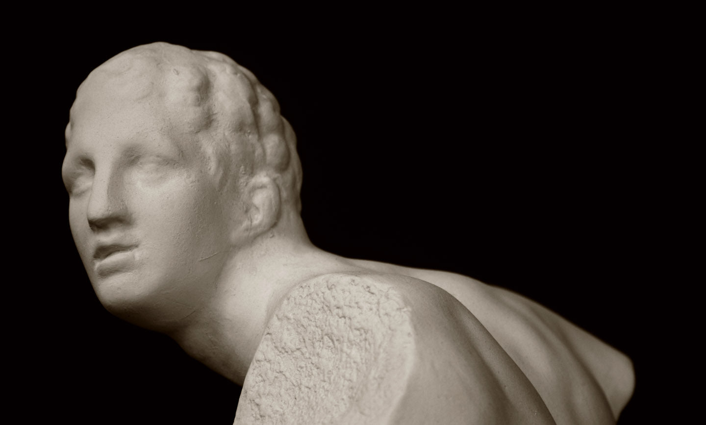 closeup photo of white plaster cast sculpture of male figure in active pose without arms or lower legs on a base against black background