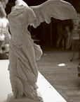 photo of plaster cast sculpture of winged, headless female figure with flowing drapery on a white shelf with wood floor visible and other plaster casts visible in background
