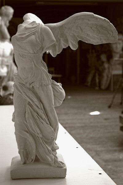 photo of plaster cast sculpture of winged, headless female figure with flowing drapery on a white shelf with wood floor visible and other plaster casts visible in background