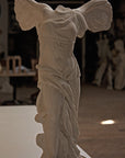 photo of plaster cast sculpture of winged, headless female figure with flowing drapery on a white shelf with wood floor visible and other plaster casts visible in background