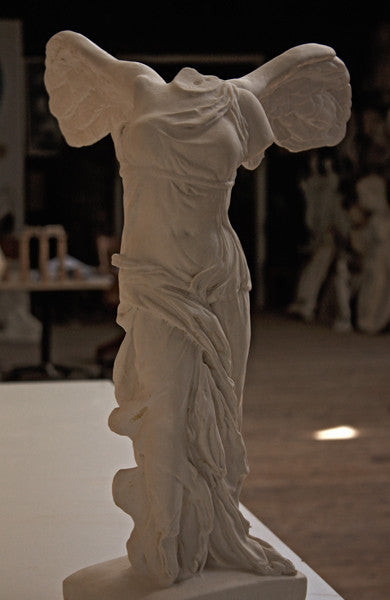 photo of plaster cast sculpture of winged, headless female figure with flowing drapery on a white shelf with wood floor visible and other plaster casts visible in background