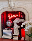 photo of white plaster cast sculpture of winged, headless female figure with flowing drapery in a white, ornate fireplace displayed on a stack of books with another stack of books beside it, disco balls hanging, red neon sign of the word dream hanging, and flowers to the right outside the fireplace