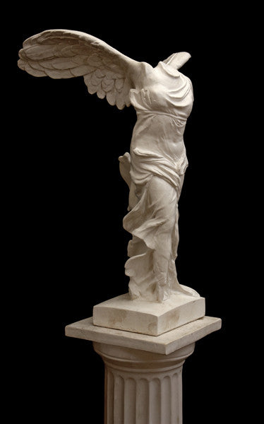 photo of plaster cast sculpture of winged, headless female figure with flowing drapery on top of a Doric-column pedestal on a black background