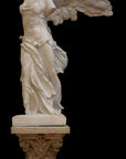 photo of plaster cast sculpture of winged, headless female figure with flowing drapery on top of an ornate, floral pedestal on a black background