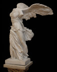 photo of plaster cast sculpture of winged, headless female figure with flowing drapery on top of an ornate, floral pedestal on a black background