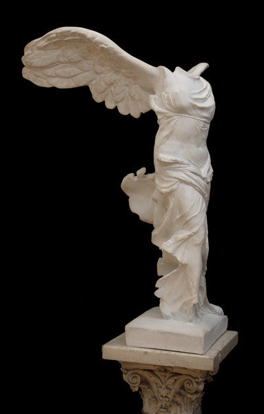 photo of plaster cast sculpture of winged, headless female figure with flowing drapery on top of an ornate, floral pedestal on a black background