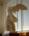 photo of plaster cast sculpture of winged, headless female figure with flowing drapery on top of a ribbed, square, wooden pedestal with white paneled walls behind