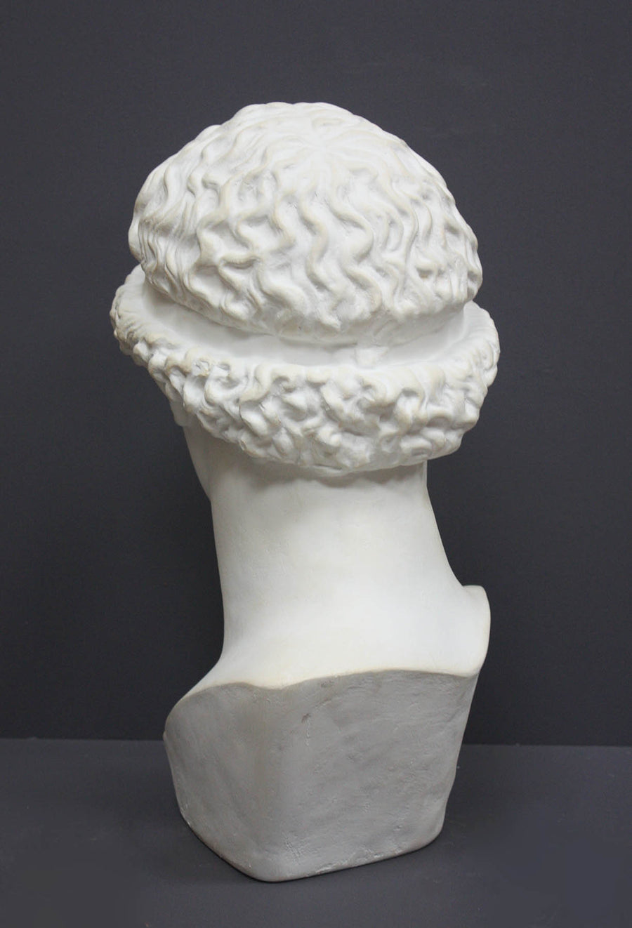 back view photo of white plaster cast sculpture bust of female, namely the goddess Athena, with short, curly hair and headband on gray background