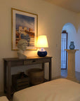 plaster cast fluted pedestal with blue vase on top near arched doorway and sculpture bust of female figure, namely the goddess Athena, with short, curly hair and headband on a wooden table next to blue lamp and in front of painting in white-walled room