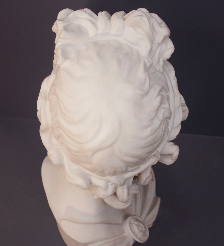 top view photo of white plaster cast sculpture bust of man, namely the god Apollo, with hair piled high in the front and a broach near his neck holding robes on a gray background