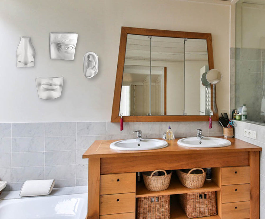 Photo of a bathroom double sink and mirror with a white wall and four plaster sculptures of David's eye, ear, nose, and mouth hanging on the wall