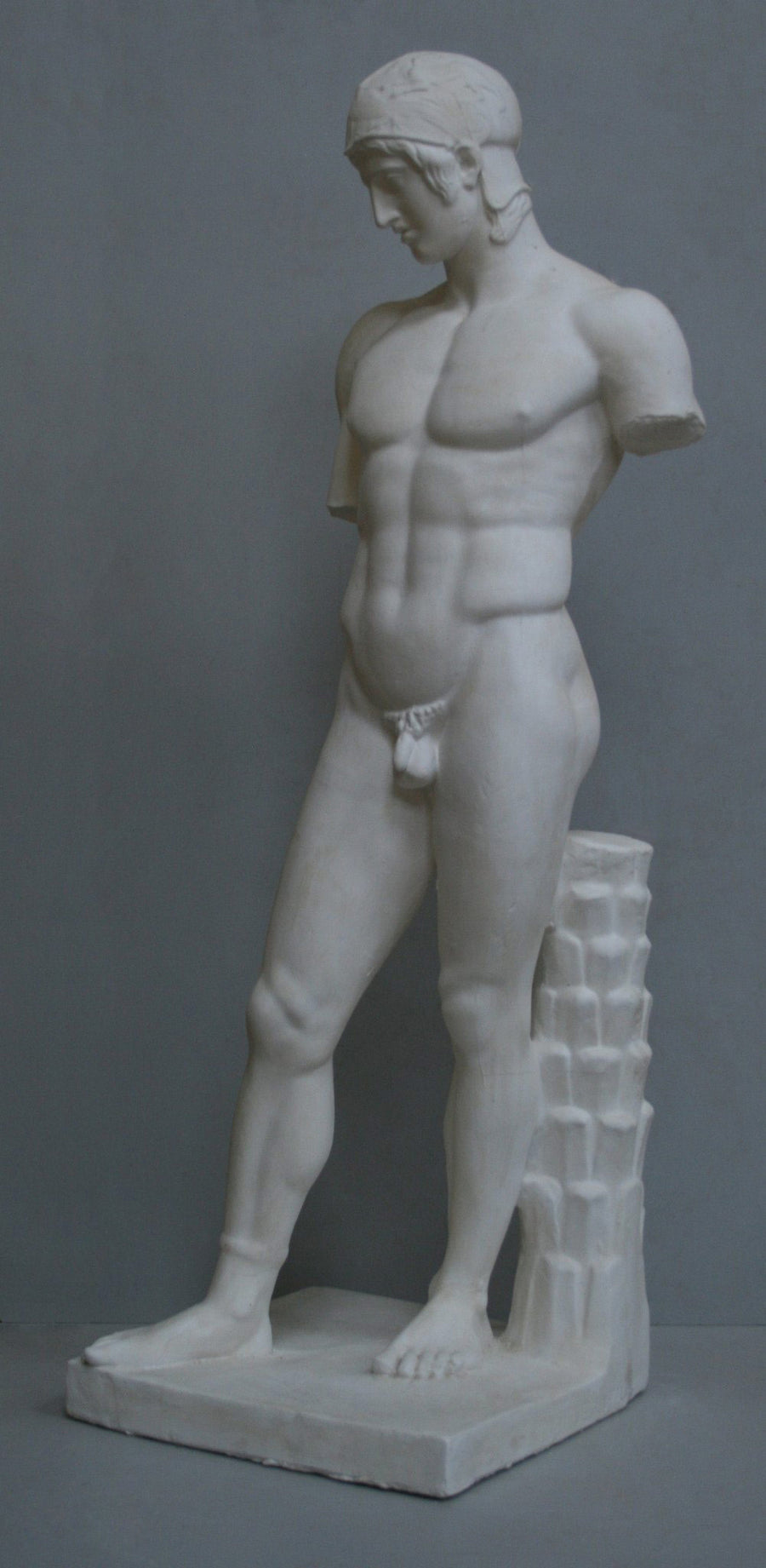 photo of plaster cast of sculpture of nude male, namely the god Ares, without arms and wearing a helmet with a supporting tree trunk, on a gray background