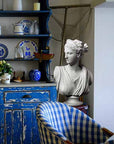 Photo of white plaster cast of sculpture bust of female goddess Diana with drapery and crown and up-do in a kitchen with blue hutch and chair