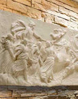 photo of a plaster cast sculpture relief of a woman, namely the goddess Aurora, flying and leading a chariot with a man pulled by horses towards the right while a cherub and other women fill the scene, on a tan brick wall