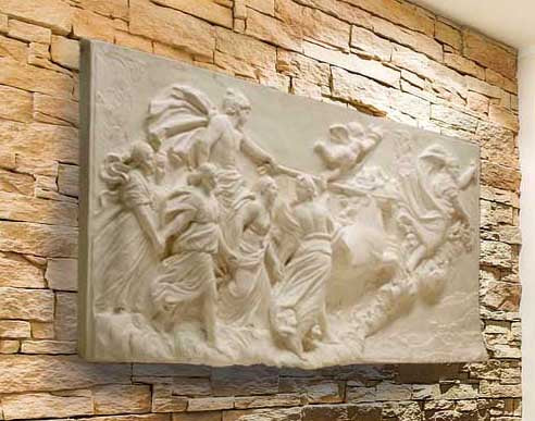 photo of a plaster cast sculpture relief of a woman, namely the goddess Aurora, flying and leading a chariot with a man pulled by horses towards the right while a cherub and other women fill the scene, on a tan brick wall