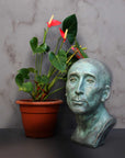 photo of plaster cast sculpture bust of man, namely Niccolo da Uzzano, in verdigris patina next to plant with pink flowers in brown pot in front of a white marble wall and on a gray surface