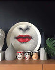 Photo of still life with three cups with faces, a dish with red lips, a plant and a vase and a plaster cast of a bust of Meleager on a shelf