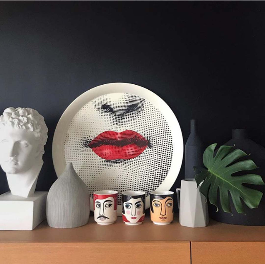 Photo of still life with three cups with faces, a dish with red lips, a plant and a vase and a plaster cast of a bust of Meleager on a shelf