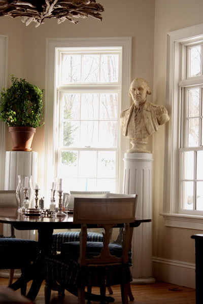 Photo of off-white plaster cast bust sculpture of male in Revolutionary garb, namely George Washington, on white pedestal in front of windows in a dining room