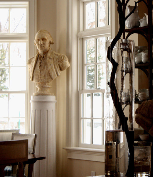Photo of off-white plaster cast bust sculpture of male in Revolutionary garb, namely George Washington, on white pedestal in front of windows in a dining room