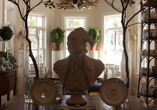 photo of plaster cast of male bust sculpture, namely Lafayette, in uniform on a table with decorative dishes and other furniture, dishes, plants, and bay windows behind